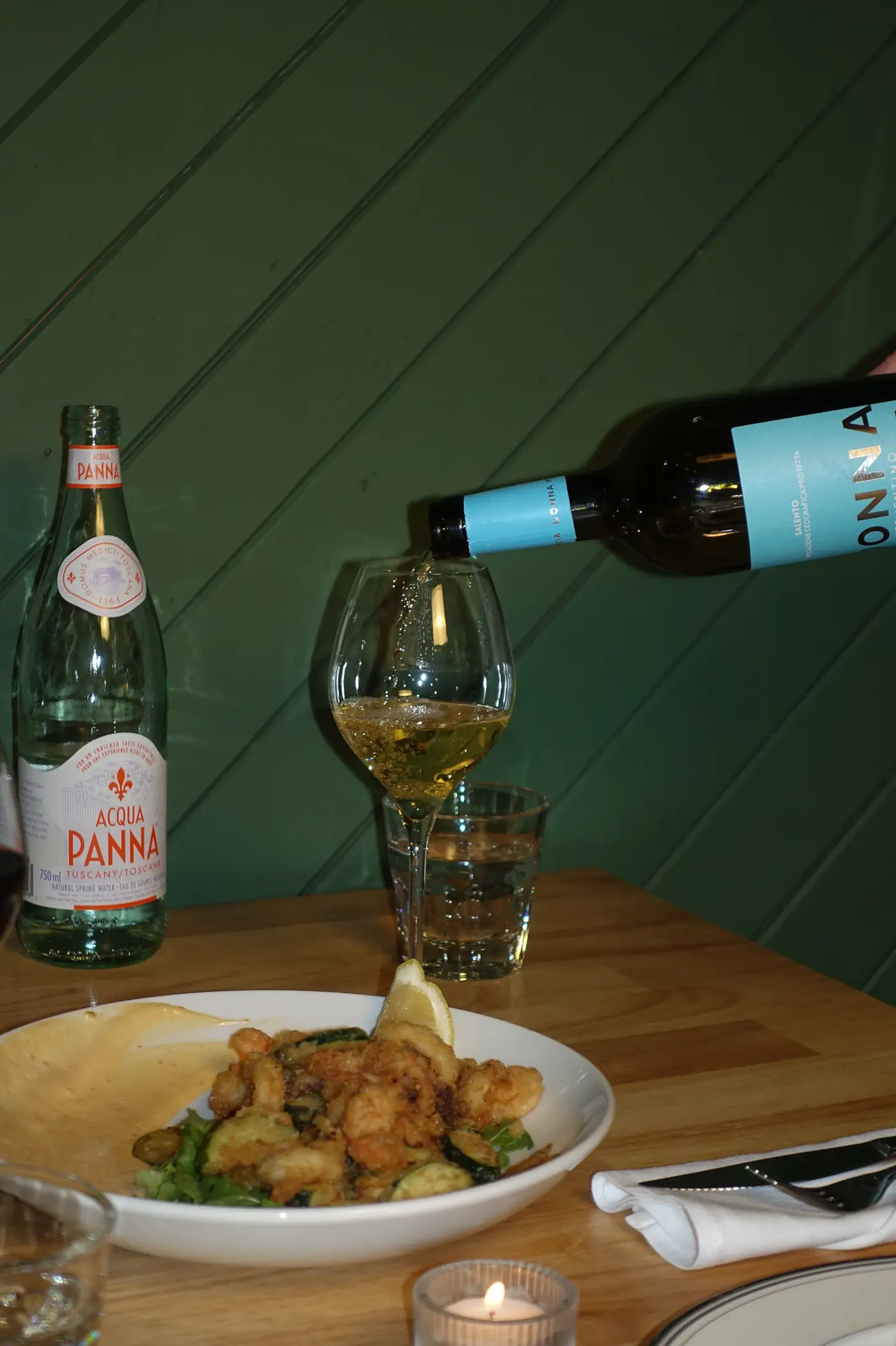 Vin blanc versé à côté d’un plat de calmars et de courgettes frits, devant un mur vert